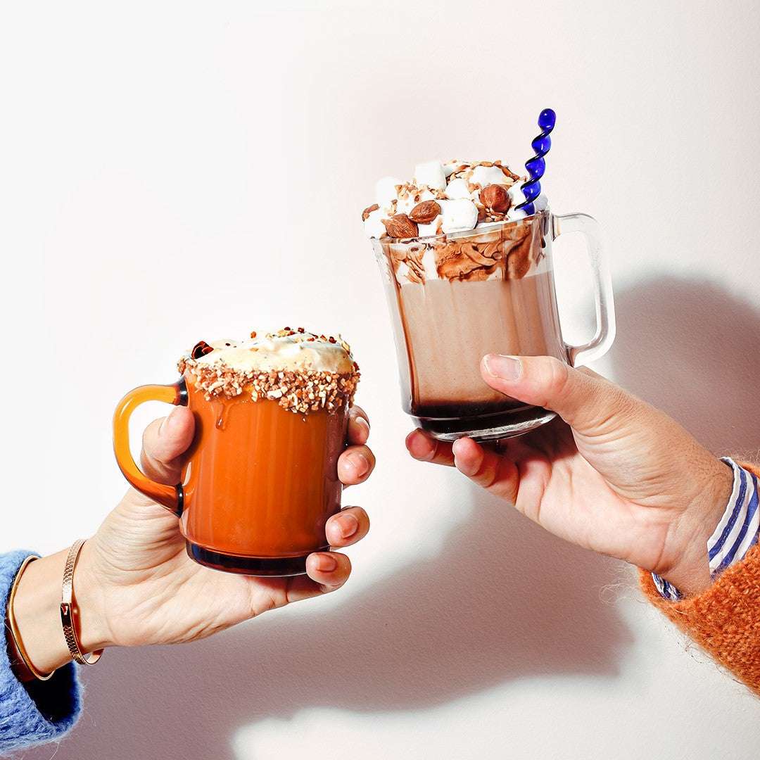 Two Duralex Versailles Stackable Glass Mugs being toasted, one in orange with toppings and the other filled with chocolate and marshmallows.