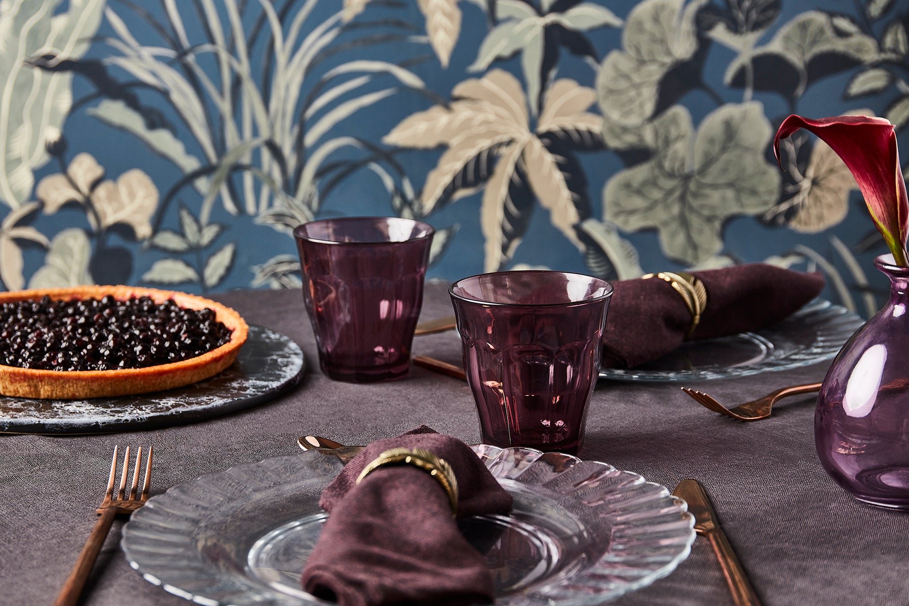 Table setting with purple glasses, napkins, and a decorative plate against a floral wallpaper background.