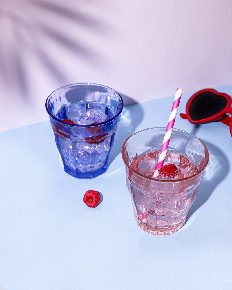 Two Le Picardie® Blush Pink Glass Tumblers by Duralex displayed alongside a blue tumbler, with raspberries and a straw on a colorful background.