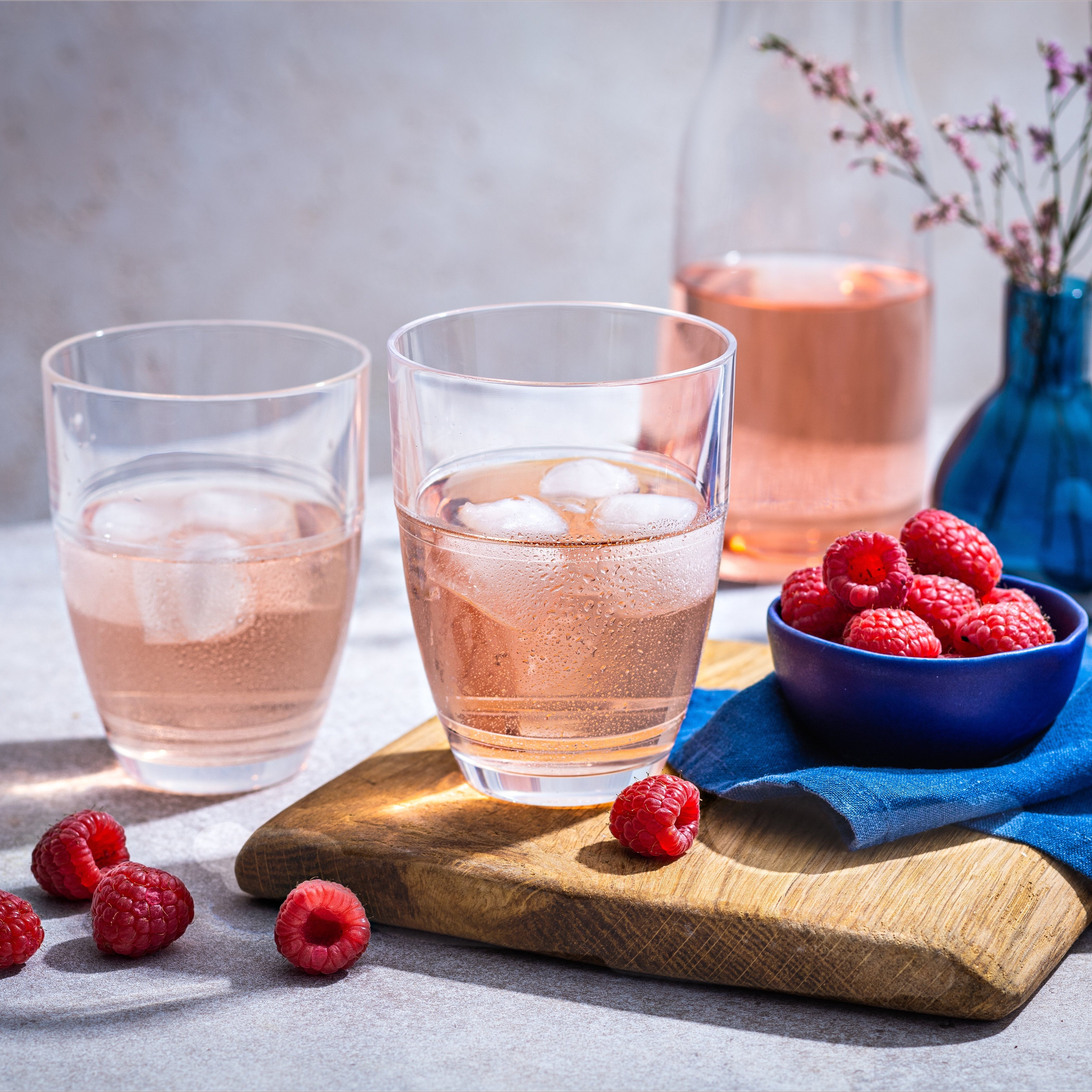 Two glasses of pink drink with ice and raspberries on a wooden board, with a blue napkin and vase in the background.